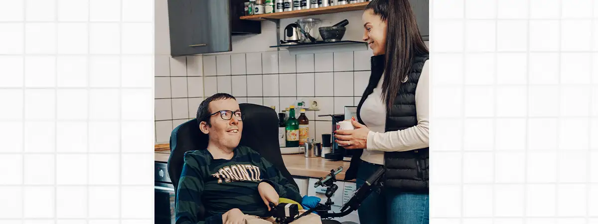 Ein Foto von Roberto, der in seinem Rollstuhl in der Küche sitzt und sich mit seiner Assistentin unterhält, die neben ihm steht.