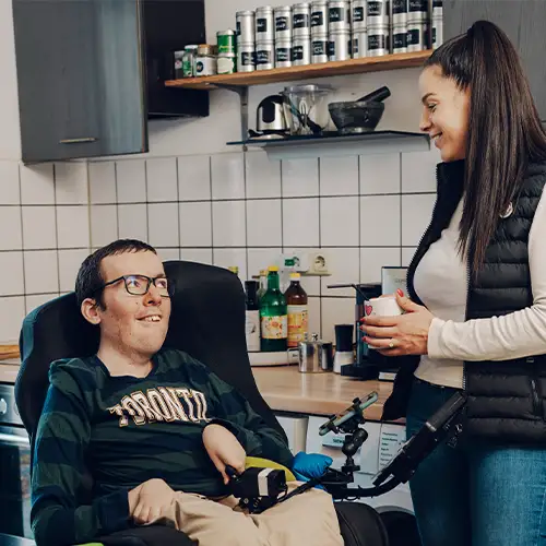 Ein Foto von Roberto, der in seinem Rollstuhl in der Küche sitzt und sich mit seiner Assistentin unterhält, die neben ihm steht.