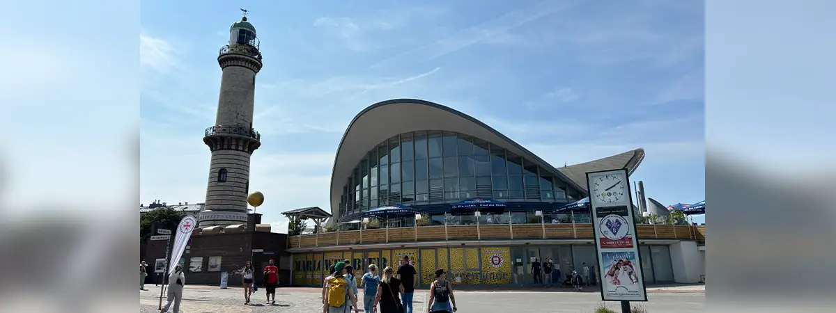 Ein Foto des Leuchtturms und der Sehenswürdigkeit „Teepott“ in Warnemünde, vor denen Menschen in Richtung der Gebäude gehen.