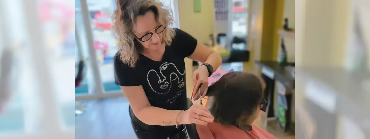 Camilla sitzt in einem Friseurstuhl und lässt sich von einer Frau die Haare schneiden. 