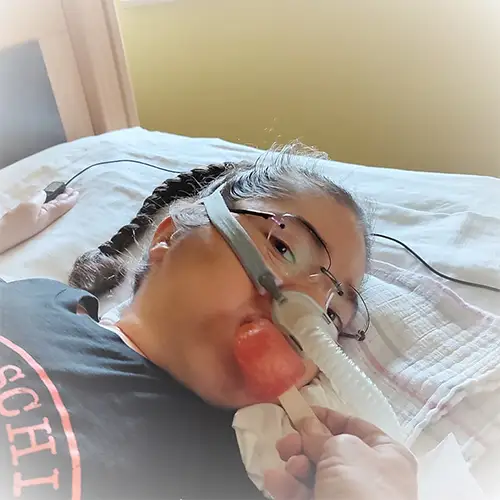 Ein Foto von Rosalie, die liegend nach vorne schaut, während sie beatmet wird und ihr ein Eis an den Mund gehalten wird.