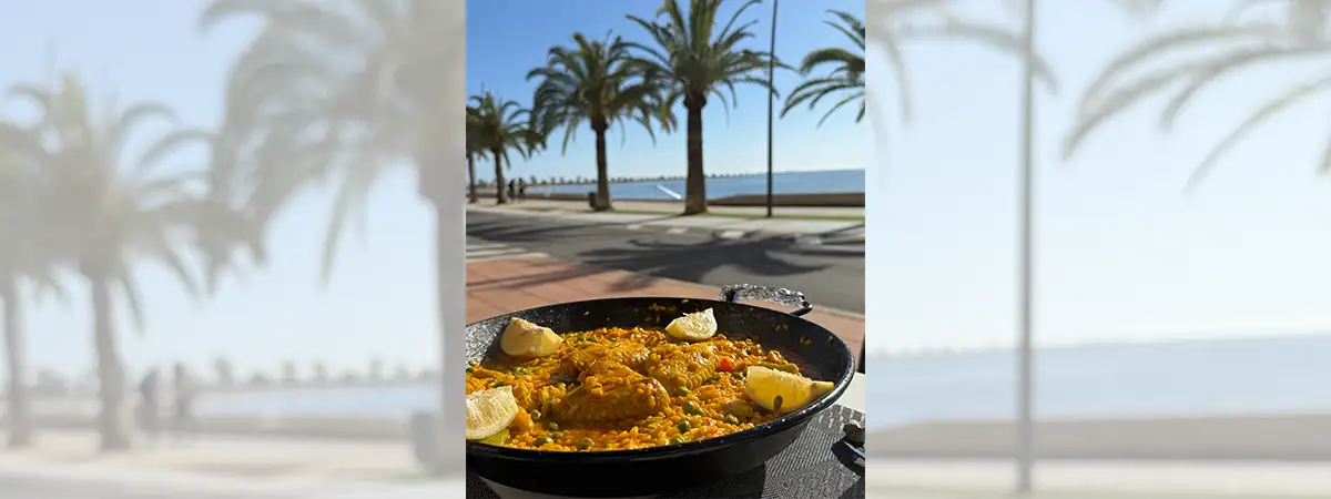 Ein Foto einer Paella in einer gusseisernen Pfanne, im Hintergrund eine Promenade mit Palmen und Blick auf das Meer.