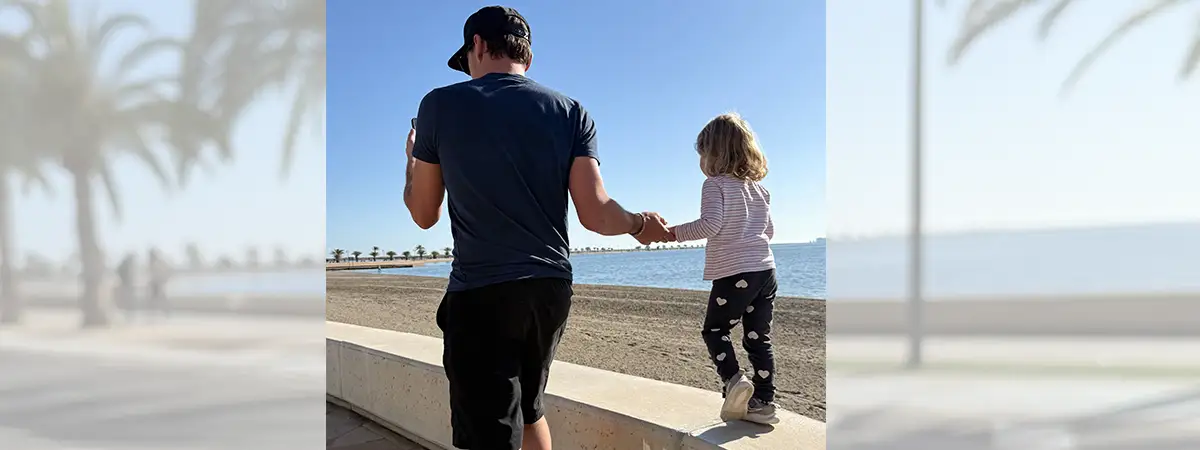 Ein Foto von Svenjas Mann und ihrer Tochter, wie sie an der Strandpromenade entlanglaufen, während das Kind auf einer Mauer balanciert.