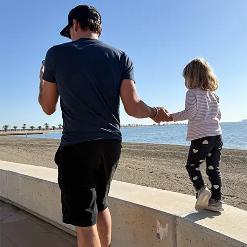 Ein Foto von Svenjas Mann und ihrer Tochter, wie sie an der Strandpromenade entlanglaufen, während das Kind auf einer Mauer balanciert.