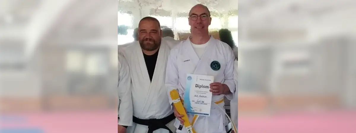 Ein Foto von Ulf, der seinen gelben Gürtel und eine Urkunde in den Händen hält, während sein Trainer und er Karateanzüge tragen und nach vorne blicken.