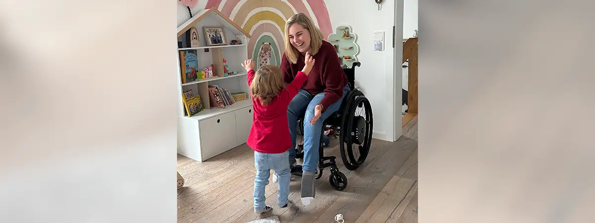 Svenja sitzt im Rollstuhl im Kinderzimmer und streckt ihrer Tochter die Arme entgegen; ihre Tochter läuft mir erhobenen Armen auf sie zu