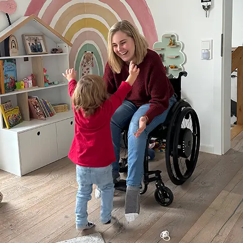 Svenja sitzt im Rollstuhl im Kinderzimmer und streckt ihrer Tochter die Arme entgegen; ihre Tochter läuft mir erhobenen Armen auf sie zu