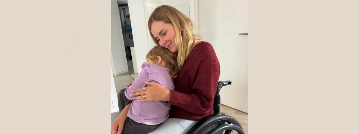 Svenja sitzt im Rollstuhl und hat ihre Tochter auf dem Schoß, welche sich an sie kuschelt