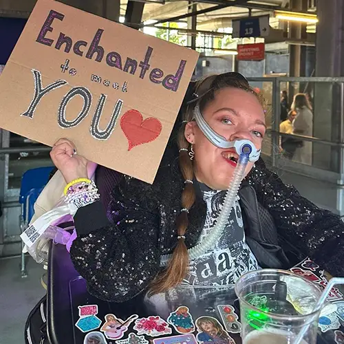 Ein Foto von Vanessa, die in ihrem mit Aufklebern verzierten Rollstuhl sitzt, ein glitzerndes Outfit trägt und ein Plakat mit der Aufschrift „Enchanted to meet you“ hochhält. 