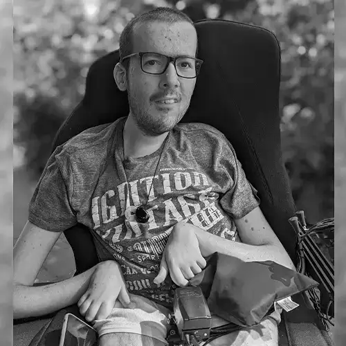 Ein schwarz-weiß Foto von Roberto, der in seinem Rollstuhl sitzt und ein T-Shirt und eine kurze Hose trägt. Im Hintergrund ist ein Busch zu sehen und die Sonne scheint. 