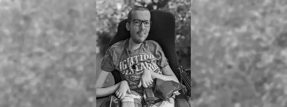 Ein schwarz-weiß Foto von Roberto, der in seinem Rollstuhl sitzt und ein T-Shirt und eine kurze Hose trägt. Im Hintergrund ist ein Busch zu sehen und die Sonne scheint. 