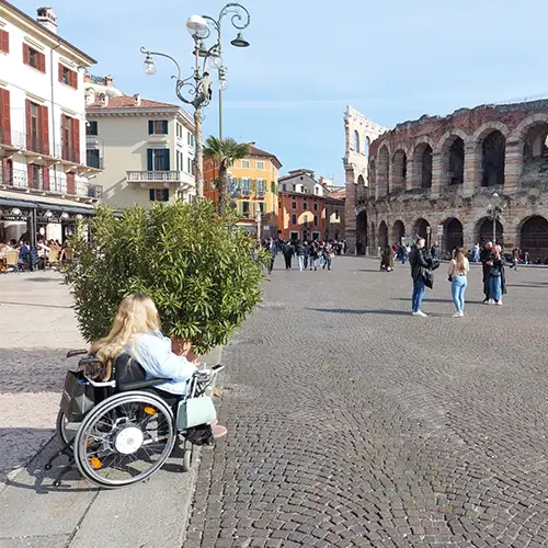 Liv sitzt im Rollstuhl und blickt auf ein Amphittheater, während sich im Hintergrund ein belebter Platz unter strahlend blauem Himmel erstreckt.