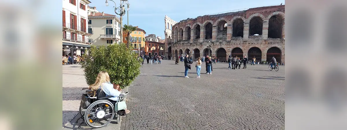 Liv sitzt im Rollstuhl und blickt auf ein Amphittheater, während sich im Hintergrund ein belebter Platz unter strahlend blauem Himmel erstreckt.