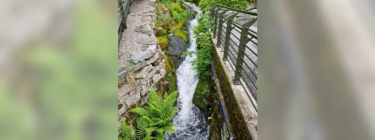 Ein Wasserfall, der zwischen Steinmauern und grünem Farn fließt - umgeben von Geländern und Brücken.