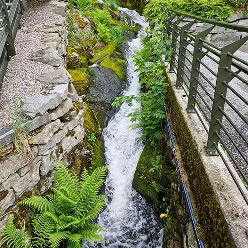 Ein Wasserfall, der zwischen Steinmauern und grünem Farn fließt - umgeben von Geländern und Brücken.