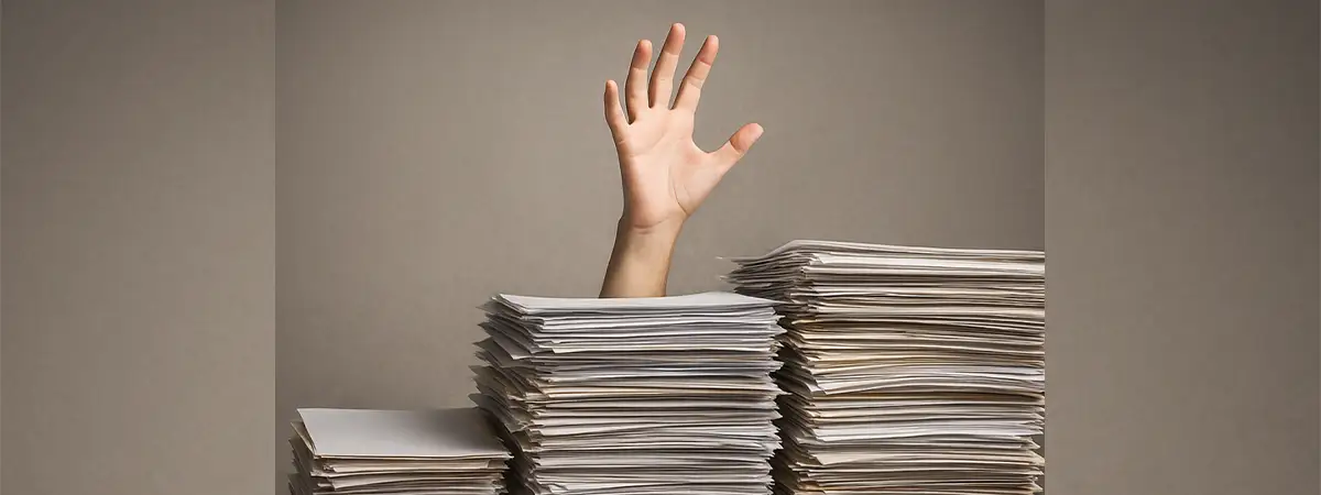 Ein Foto einer Hand, die hinter großen Papierstapeln auf einem Schreibtisch hervorragt.