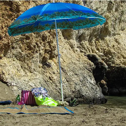 Ein blauer Sonnenschirm steht neben einem Felsen am Strand, daneben liegen eine Matte und Taschen.