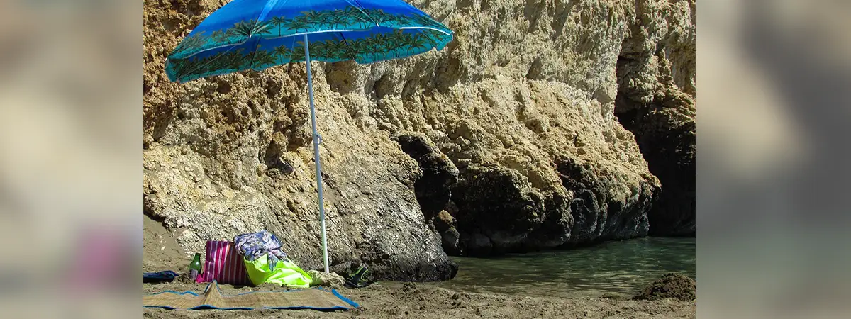Ein blauer Sonnenschirm steht neben einem Felsen am Strand, daneben liegen eine Matte und Taschen.