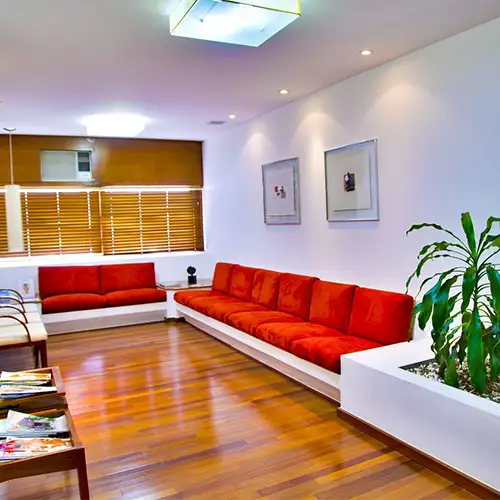 Ein Wartebereich einer Arztpraxis mit roten Sitzbänken, Holzboden, Pflanzen und Zeitschriften.