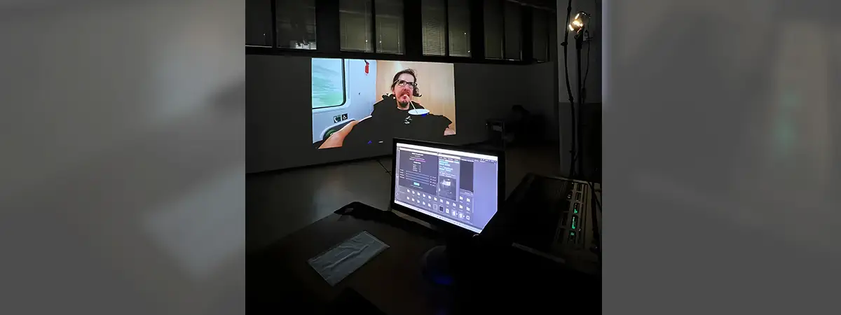 Christian ist auf einer Leinwand zu sehen und sitzt in seinem eRollstuhl. Im Bild davor steht ein Laptop auff einem dunklen Schreibtisch. 