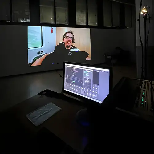 Christian ist auf einer Leinwand zu sehen und sitzt in seinem eRollstuhl. Im Bild davor steht ein Laptop auff einem dunklen Schreibtisch. 