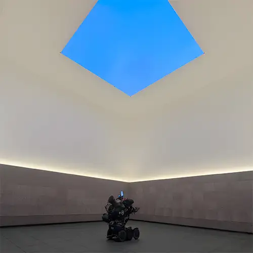 Christian sitzt in seinem eRollstuhl und betrachtet eine blaue Kunstinstallation an der Decke eines Museums. 