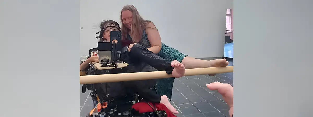 Christian sitzt in seinem eRollstuhl neben ihm lehnt seine Partnerin Grit auf ihm. Beide heben jeweils einen Fuß auf eine Ballettstange vor einem Spiegel.