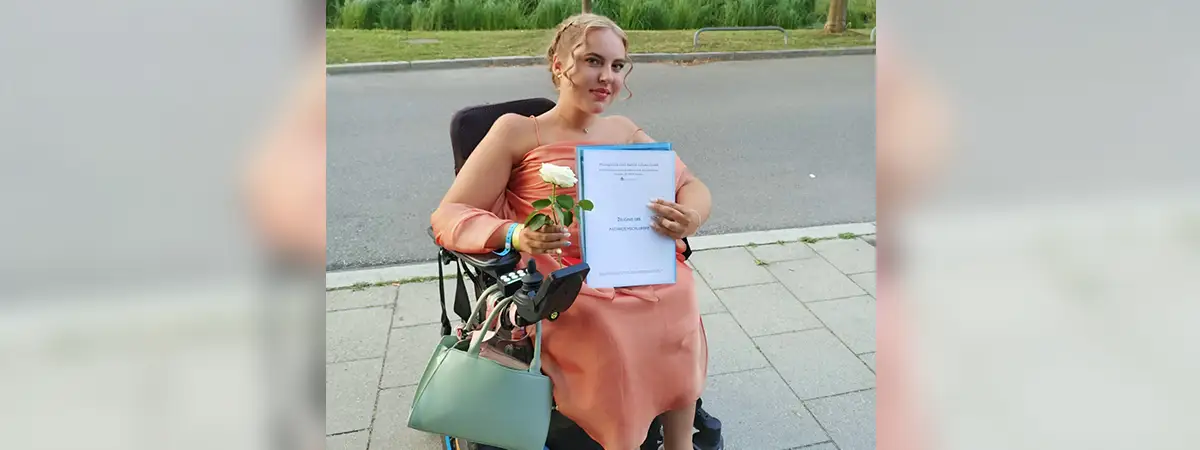 Eine junge Frau mit SMA im Rollstuhl hält stolz ihr Abi-Zeugnis in der Hand.