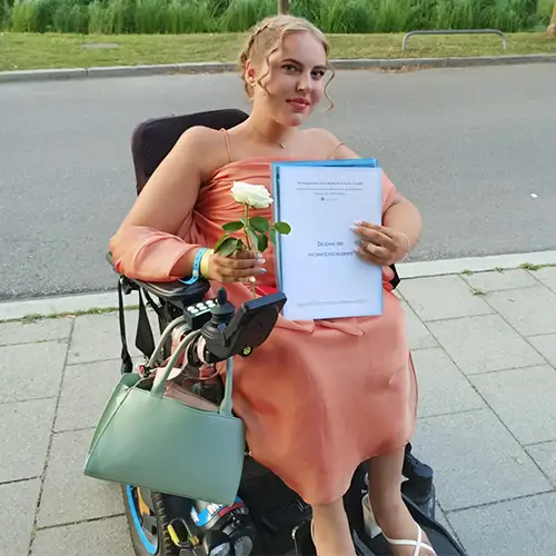 Eine junge Frau mit SMA im Rollstuhl hält stolz ihr Abi-Zeugnis in der Hand.