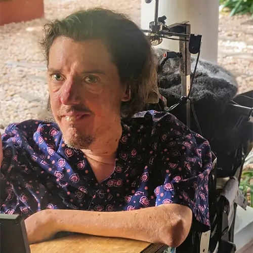 Christian lächelt aus seinem Rollstuhl in die Kamera und trägt dabei ein lilafarbenes Hemd.