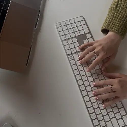 Ein Foto von oben von Amelies Händen beim Tippen auf einer Tastatur neben ihrem Laptop.