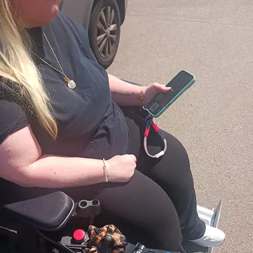 Sandra sitzt in ihrem E-Rollstuhl vor einem Fahrzeug und hält ein Handy in der Hand.