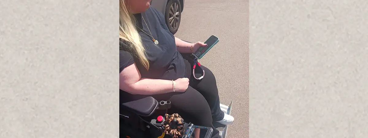Sandra sitzt in ihrem E-Rollstuhl vor einem Fahrzeug und hält ein Handy in der Hand.