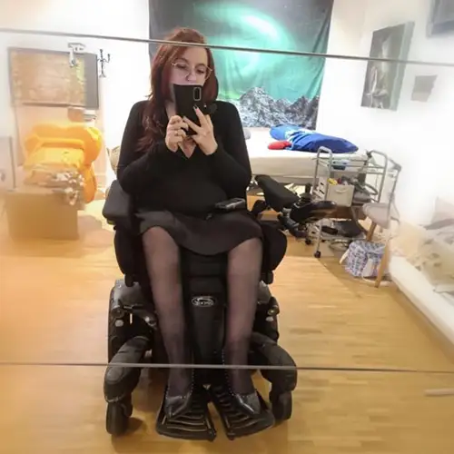 Lys sitzt in ihrem E-Rollstuhl vor einem Spiegel in ihrem Zimmer