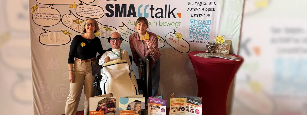 Austausch am SMAlltalk-Informationsstand beim SMA-Symposium: Bastian und zwei Vertreterinnen von Biogen am Biogen Stand. Auf dem Stand liegen Informationsmaterialien aus, die Rückwand zeigt das SMAlltalk-Logo und handschriftlich ausgefüllte Sprechblasen mit Beiträgen der Community.