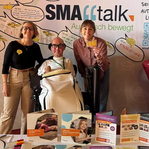 Austausch am SMAlltalk-Informationsstand beim SMA-Symposium: Bastian und zwei Vertreterinnen von Biogen am Biogen Stand. Auf dem Stand liegen Informationsmaterialien aus, die Rückwand zeigt das SMAlltalk-Logo und handschriftlich ausgefüllte Sprechblasen mit Beiträgen der Community.