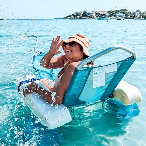 Alt text: Kim ist mit einem Rollstuhl im Meer und winkt lächelnd in die Kamera, im Hintergrund sieht man andere Menschen und weiter entfernt eine Küste mit Booten und Häusern.