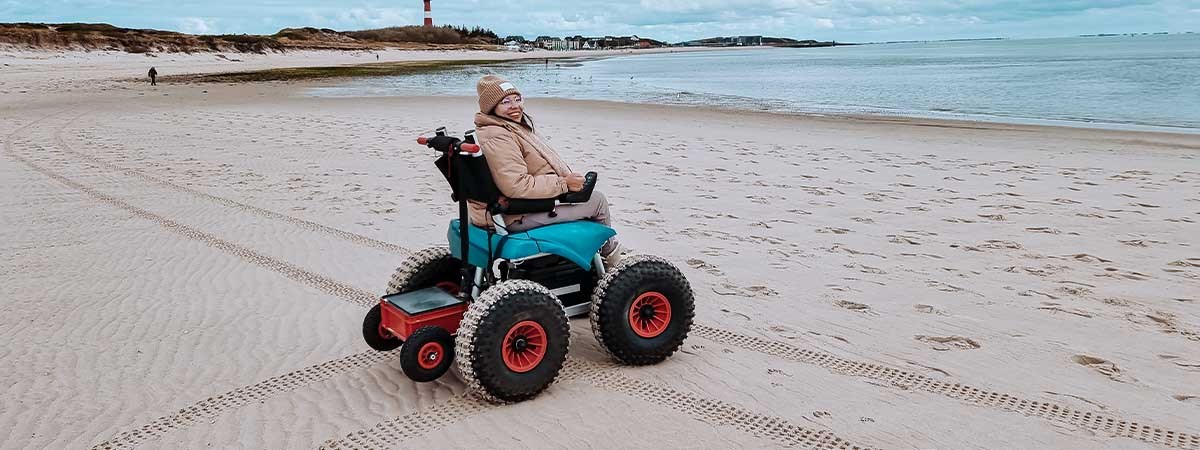 Kim ist in warmer Kleidung mit einem E-Strandrollstuhl auf dem Sand unterwegs, im Hintergrund sieht man das Meer und einen Leuchtturm.