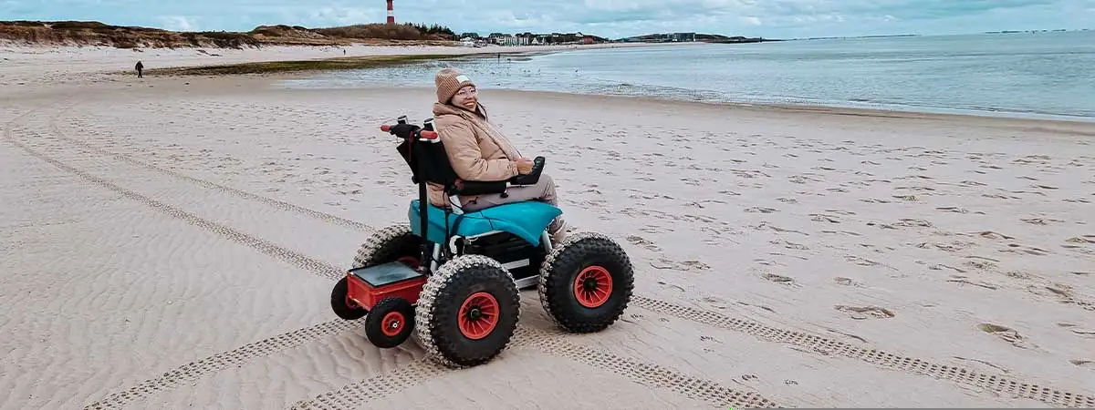 Kim ist in warmer Kleidung mit einem E-Strandrollstuhl auf dem Sand unterwegs, im Hintergrund sieht man das Meer und einen Leuchtturm.