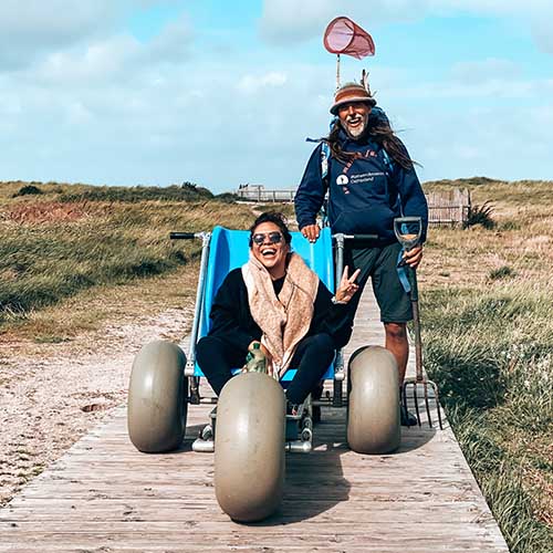 Kim sitzt lächelnd in ihrem Rollstuhl am Strand. Neben ihr ist eine Anlage mit einem auf Schienen angebrachten Sitz, die ins Meer führt. Im Hintergrund sieht man das Meer und viele Menschen, im Vordergrund ein Holzschild mit einem Zeichen für Rollstühle.