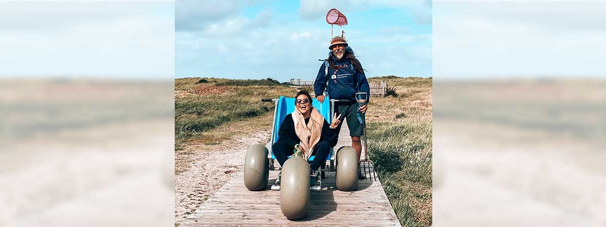 Kim sitzt lächelnd in ihrem Rollstuhl am Strand. Neben ihr ist eine Anlage mit einem auf Schienen angebrachten Sitz, die ins Meer führt. Im Hintergrund sieht man das Meer und viele Menschen, im Vordergrund ein Holzschild mit einem Zeichen für Rollstühle.