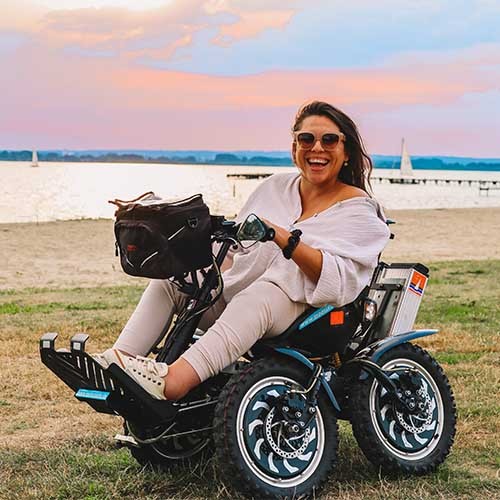 Eine lachende Frau im Rollstuhl auf einer Wiese vor einem See. 