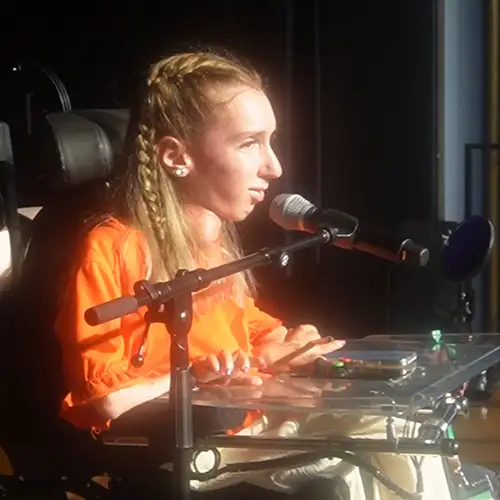 Ein Bild, das Autorin Celina von der Seite in ihrem elektrischen Rollstuhl auf einer Bühne zeigt. Vor ihr ist ein Mikrofon in einem Ständer.