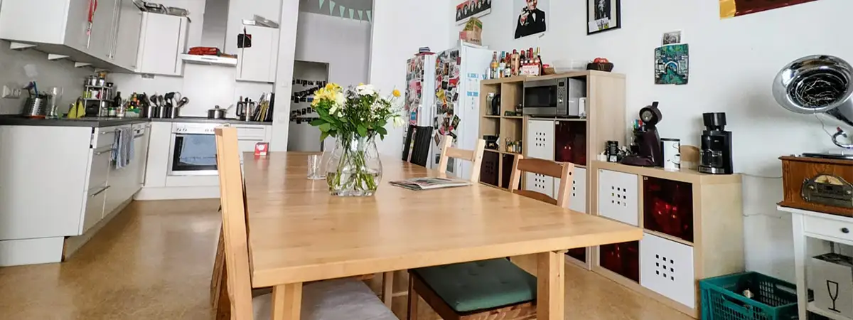 Ein Bild aus Christians WG-Küche, in deren Mitte ein Holztisch mit einer Blumenvase steht. Die Wände sind dekoriert mit Bildern, Fotos und Girlanden.