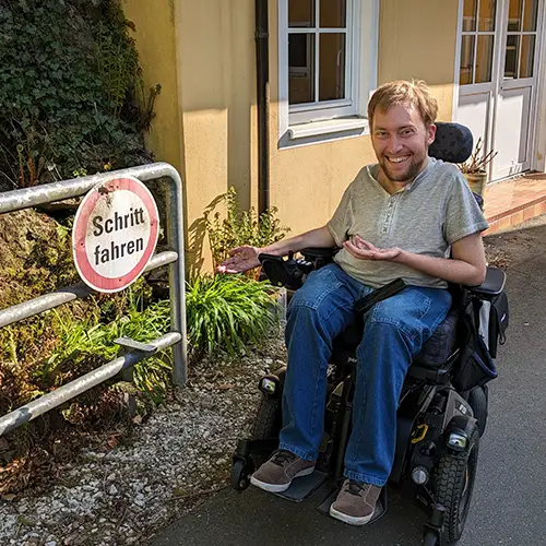Autor Christian befindet sich in seinem elektrischen Rollstuhl auf einer Straße und deutet lachend auf ein Verkehrsschild mit der Aufschrift „Schritt fahren“.