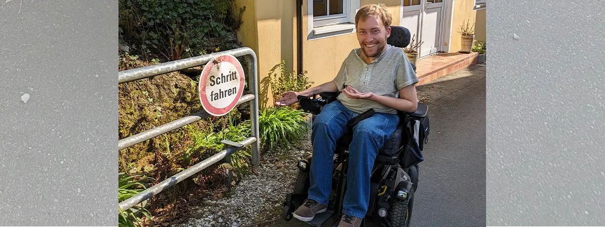 Autor Christian befindet sich in seinem elektrischen Rollstuhl auf einer Straße und deutet lachend auf ein Verkehrsschild mit der Aufschrift „Schritt fahren“.
