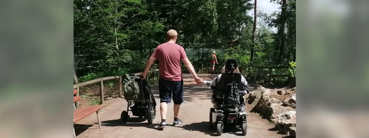 Sandras Mann schiebt mit der linken Hand einen Kinderwagen, an der rechten Hand hält er Sandra, die in ihrem Rollstuhl sitzt
