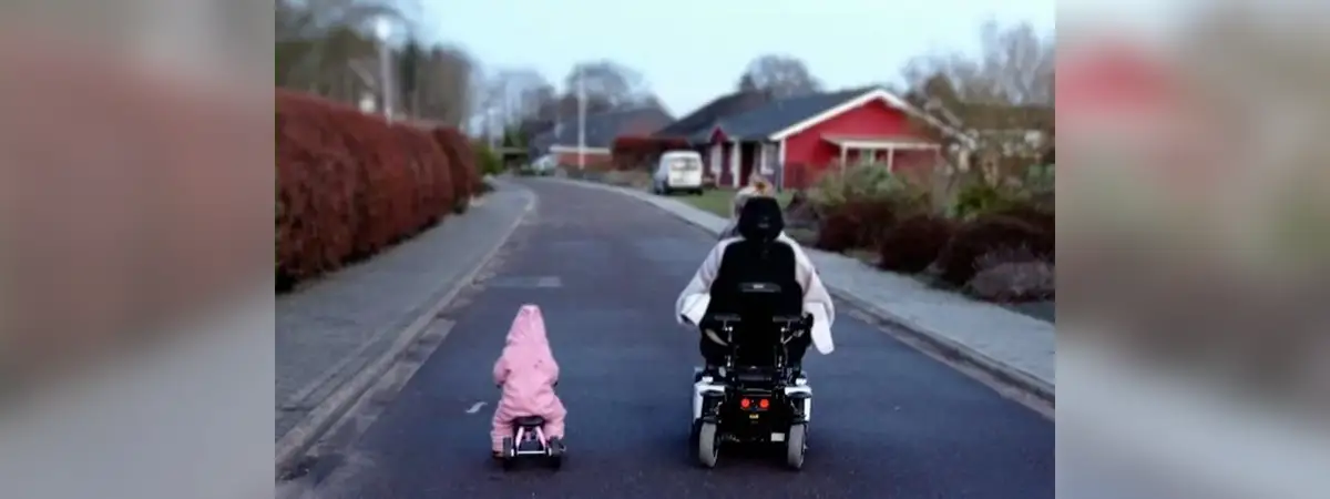 Sandra und ihre kleine Tochter von hinten wie sie eine Straße entlang fahren, Sandra im Rollstuhl, die Tochter auf dem Laufrad