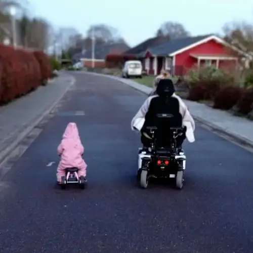 Sandra und ihre kleine Tochter von hinten wie sie eine Straße entlang fahren, Sandra im Rollstuhl, die Tochter auf dem Laufrad