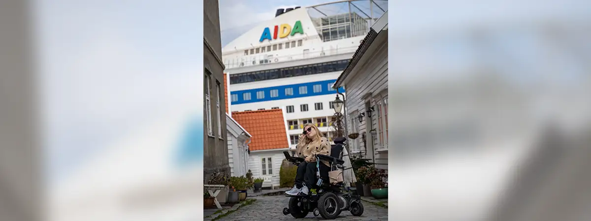 Ein Foto von Lilli im Elektrorollstuhl, während hinter ihr ein AIDA-Kreuzfahrtschiff ist.